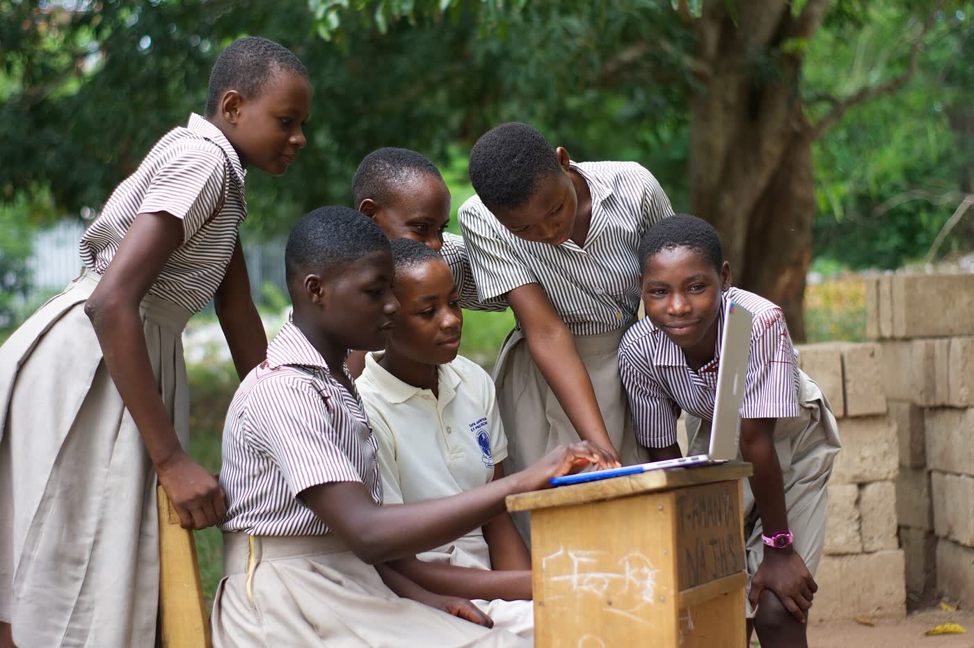 Five students in school uniform gathered around a laptop outdoors